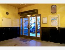 Eingangsbereich mit gelb gestrichenen Wänden, teils schwarz abgesetzt. Im Fokus stehen eine gläserne Doppeltür mit dekorativen Metallkreisen und ein Türgriff im Ringdesign. Links befindet sich eine weiße Tür, rechts ein Aushang mit Plakat und Grundriss.