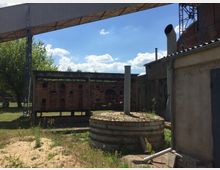 Industriegelände mit Backsteinmauer, altem Beton- und Metallbau sowie einem Zylinder aus Beton in der Mitte. Im Hintergrund sind Bäume, Wiesen und blauer Himmel mit Wolken zu sehen.
