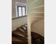 Gewundene Steintreppe in einem historischen Gebäude mit weiß verputzten Wänden. Im Hintergrund ist ein großes Fenster mit schwarzem Rahmen zu sehen, das natürliches Licht hereinlässt.