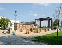 Ein Platz mit gepflastertem Weg, grünem Rasen und einer offenen Freifläche mit gestapelten Holzpaletten. Im Hintergrund ein rotes Backsteingebäude und eine moderne schwarze Metallkonstruktion, rechts angrenzend Bäume und Autos.