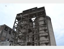 Industrieanlage mit mehrstöckigen Betonsilos und freiliegendem Trägergerüst, umgeben von einem alten Fabrikgebäude mit zerbrochenen Fenstern; im Hintergrund ein bewölkter Himmel mit einem Flugzeug in der Ferne.