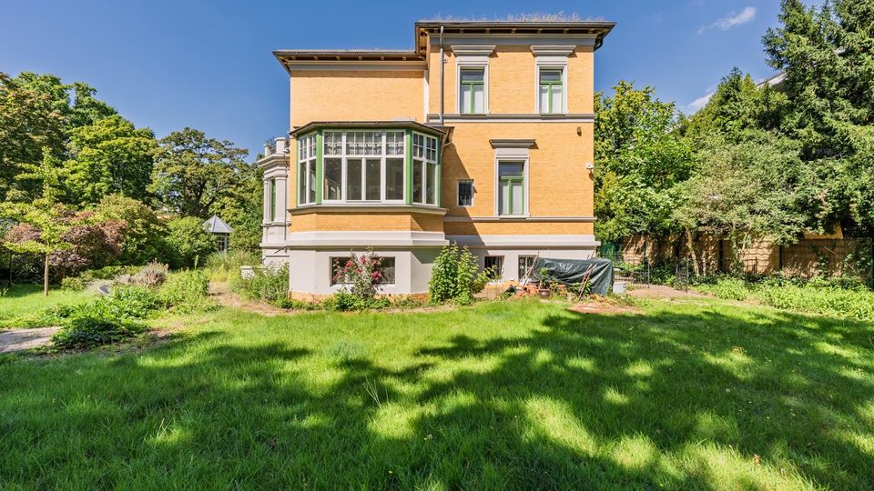 Mehrstöckige gelbe Backsteinvilla mit großen Fenstern und verglastem Erker, umgeben von einem weitläufigen Garten mit Rasenfläche, Sträuchern und hohen Bäumen. Das Gebäude hat einen hell abgesetzten Sockel und steht vor blauem Himmel in einer grünen, parkähnlichen Umgebung.