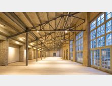 Großer, offener Industrie-Innenraum mit Ziegelwänden, freiliegender Stahlkonstruktion an der Decke und großen Fensterfronten, die Tageslicht hereinlassen. Der Boden besteht aus hellen Holzdielen, und dezente Beleuchtung betont die architektonischen Details.