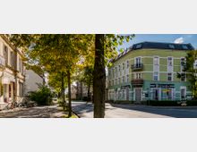 Straßenszene mit einem grün gestrichenen, historischen Gebäude mit weißen Verzierungen und Balkonen auf der rechten Seite, das als Restaurant und Musikbar dient. Links säumen Bäume und weitere Altbauten den Gehweg, dahinter verläuft eine ruhige, sonnige Straße.