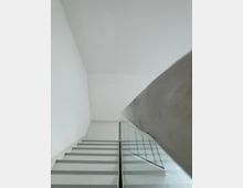 Eine minimalistische, gerade Treppe in einem weißen Innenraum, mit einem silbernen Handlauf und transparenten Glaswänden als Geländer. Die Umgebung wirkt schlicht und modern mit klaren Linien und neutralen Farben.