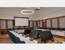 Moderner Konferenzraum mit langen, weißen Tischen in U-Form, ausgestattet mit Mikrofonen und Monitoren. Im Hintergrund befinden sich eine Projektionsfläche, Holzwandpaneele und eine Tür, die von schallisolierten Elementen umgeben ist; eine kreisförmige Leuchte hängt von der Decke.