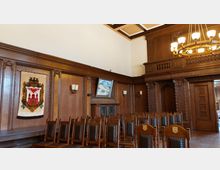 Innenraum eines historischen Sitzungssaals mit holzgetäfelten Wänden, verzierten Schnitzereien und einer großen Deckenlampe. Mehrere Reihen hölzerner, gepolsterter Stühle stehen vor einer Wand mit einem Wappen-Teppich und einem Gemälde.