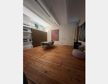 Ein Raum mit Holzdielenboden, einem rechteckigen Tisch mit Stühlen in der Mitte und moderner Einrichtung. Links befinden sich Regale mit Weinflaschen, im Hintergrund eine braune Schrankwand, darüber moderne Sessel, und an der Wand hängt ein farbiges abstraktes Gemälde.