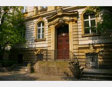 Fassade eines historischen Gebäudes mit hellen Natursteinen und dekorativen Details. Der Eingang ist durch ein hölzernes Doppeltor und einen verzierten Türbogen mit der Inschrift "Amtsgericht" hervorgehoben; seitlich führen kurze Treppen mit schmiedeeisernem Geländer hinauf.