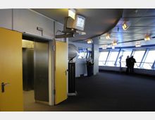 Innenansicht eines Aussichtsdecks mit gelben Türen, einem offenen Fahrstuhl, großen Fenstern mit Blick nach draußen und einem Teleskop. Der Raum ist modern gestaltet, mit einer abgehängten Decke und punktueller Beleuchtung sowie vereinzelter Dekoration wie Blumen und einem Globus.
