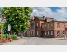 Backsteinbau eines historischen Bahnhofsgebäudes mit Satteldach und weißen Fenstern, umgeben von Bäumen. Im Vordergrund befinden sich eine Bushaltestelle mit Schild "ZOB", rote Sitzbänke und Straßenlaternen im gleichen Farbton.