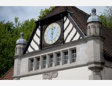 Fachwerkfassade eines historischen Gebäudes mit einem großen, runden Zifferblatt in der Giebelmitte. Die Fassade ist von Holz- und Steinelementen geprägt, flankiert von Türmen mit kleinen Kuppeln, umgeben von dichter Vegetation.