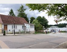 Straßenszene in einer ländlichen Gegend mit einem kleinen weiß-grauen Wohnhaus mit Satteldach im Vordergrund. Im Hintergrund befindet sich ein See mit einem Anleger, einem weißen Boot und umliegenden Bäumen.