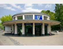 Runder Eingangsbereich des U-Bahnhofs „Krumme Lanke“ in Berlin, mit hellem Putz, Glasfenstern und einem blauen U-Bahn-Schild. Im Vordergrund ein gepflasterter Platz, links ein geschlossener Kiosk, rechts Fahrradständer und Bäume im Hintergrund.