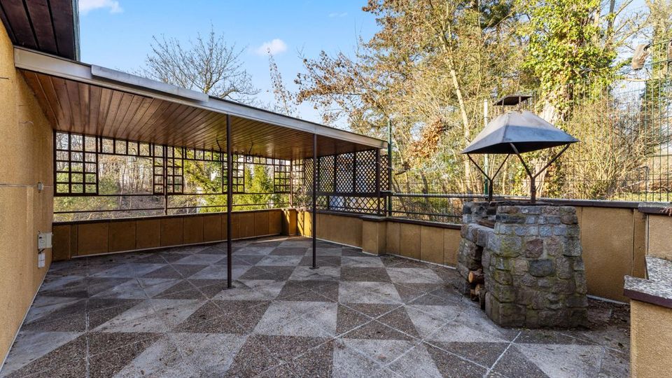Überdachte Terrasse mit gemustertem Steinboden, Holzdecke und Metallstützen, umgeben von niedrigen Wänden und Gitterelementen. Rechts steht ein gemauerter Grill- oder Kaminaufbau mit Metallhaube, dahinter Bäume und ein Zaun im Außenbereich.