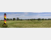 Grüne Flusslandschaft mit Wiesen, vereinzelten Bäumen und einem rot-schwarz-gelb gestreiften Grenzpfosten im Vordergrund; im Hintergrund ein Fluss und ein weiter Himmel mit leichter Bewölkung.