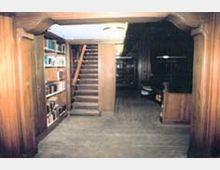 Ein Raum mit Holzverkleidung, einer zentralen Holztreppe, die nach oben führt, sowie Bücherregalen an den Wänden. Der Boden ist ebenfalls aus Holz, und die Beleuchtung ist gedämpft.