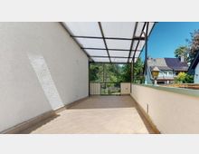 Überdachte Terrasse mit Fliesenboden, weißer Wand und einer transparenten Kunststoffdachkonstruktion. Im Hintergrund sind ein Garten und benachbarte Wohnhäuser mit Satteldächern zu sehen.