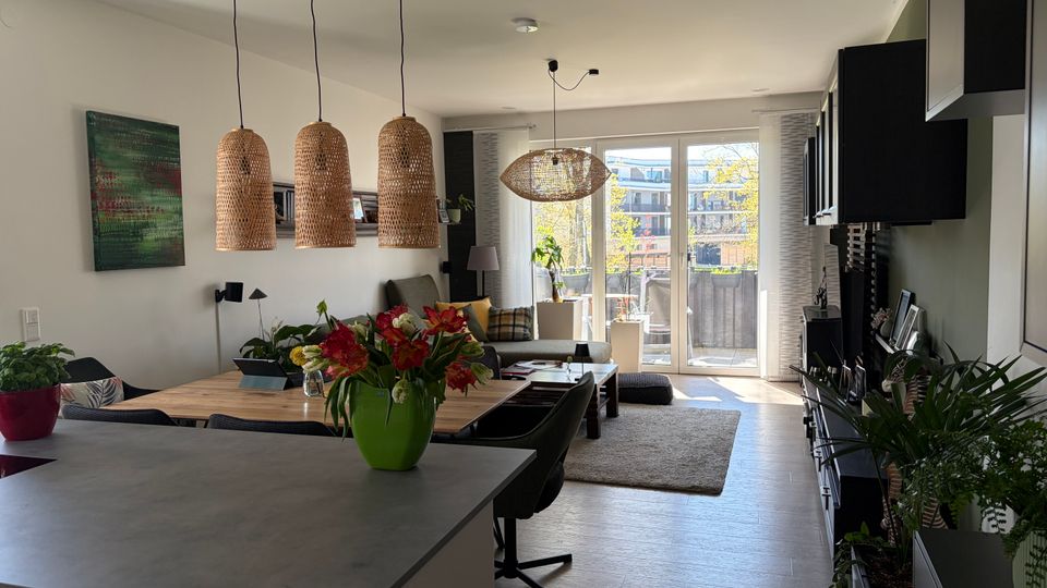 Offener Wohn- und Essbereich mit Kücheninsel im Vordergrund, Esstisch und Sofa; über dem Tisch hängen drei geflochtene Pendelleuchten. Große bodentiefe Glastüren führen auf einen Balkon, rechts eine dunkle Einbauwand mit Stauraum, dazu Pflanzen und ein Teppich auf hellem Boden.