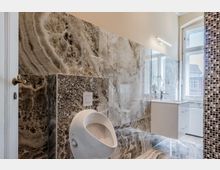 Modernes Badezimmer mit großformatigen, marmorierten Steinfliesen an Wänden und Boden, Urinal und wandhängendem Waschtisch. Rechts sorgt ein großes Fenster für Tageslicht; eine Mosaikfliesenwand und eine Leuchte sind sichtbar.