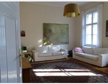 Wohnzimmer mit hellbeigen Wänden, zwei weißen Sofas, einem großen Teppich auf Holzboden und einer goldfarbenen Hängelampe. Ein großes Fenster mit weißen Vorhängen sowie ein Gemälde hängen an den Wänden.