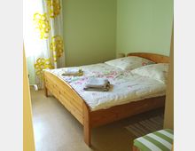 Ein kleines Schlafzimmer mit hellgrünen Wänden, einem Holzbett mit weißer Bettwäsche samt buntem Muster und gefalteten Handtüchern darauf. An der Seite ein Fenster mit gelb gemusterten Vorhängen, ein kleiner Teppich am Boden und ein gestreifter Hocker im Vordergrund.