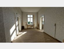 Ein leerer, renovierungsbedürftiger Raum mit abgeblättertem Putz an den Wänden, freiliegendem Holzfußboden und einem großen Fenster, das Tageslicht hereinlässt. Links und rechts führen Türen in andere Räume, deren Zustand ähnlich wirkt.