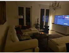 Wohnzimmer mit heller Einrichtung und gedämpfter Beleuchtung. Weiße Sofas umgeben einen Couchtisch mit brennenden Kerzen, im Hintergrund ein großer Fernseher, ein Kronleuchter, bodentiefe Fenster und ein Teppich auf hellem Boden.