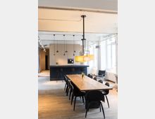 Moderner Ess- und Kochbereich mit einem langen Holztisch und 10 schwarzen Stühlen im Vordergrund. Im Hintergrund eine offene Küche mit dunkler Kücheninsel, Hängelampen, Holzfußboden und großen Fenstern, die für viel Licht sorgen.