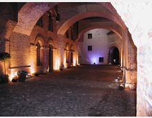 Backstein-Gewölbegang mit beleuchteten Rundbögen, gepflastertem Boden und einer beleuchteten Fassade im Hintergrund. Die Szene wirkt wie der Innenhof oder Durchgang eines historischen Gebäudes.