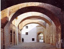 Historischer Durchgang mit Backsteinbögen und gepflastertem Boden. Links sind bogenförmige Fenster in einer Backsteinfassade, rechts eine Steinmauer, im Hintergrund ein heller Innenhof sichtbar.