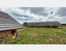 Holzhütte mit schrägem Dach links im Vordergrund und grasbedeckter Langbau mit Strohdach rechts auf einer Wiese in ländlicher Umgebung unter bewölktem Himmel. Im Hintergrund sind Bäume sichtbar.