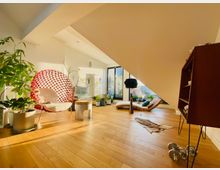 Heller Wohnraum mit Holzboden und moderner Einrichtung, darunter ein auffälliger roter Stuhl und eine Liege vor großen Fenstern. Viele Zimmerpflanzen und dekorative Elemente schaffen eine gemütliche Atmosphäre.