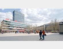 Städtischer Platz mit breiter gepflasterter Fläche, umgeben von modernen Gebäuden, darunter ein mehrstöckiges Hochhaus mit dem Mercedes-Stern auf dem Dach. Im Vordergrund gehen Menschen, während andere den Platz beleben; kahle Bäume deuten auf eine kühle Jahreszeit hin.