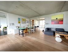 Wohnbereich in einem modernen Apartment mit offenem Grundriss, Betonwänden und hellem Holzboden. Eine graue Eckcouch, ein Esstisch mit unterschiedlich gestalteten Stühlen und große Fenster, die auf einen Balkon führen, bestimmen den Raum.