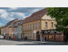 Eine Straßenansicht mit historischen Gebäuden in Pastellfarben und Backsteinfassaden, geprägt von Satteldächern und großen Fenstern. Im Vordergrund befindet sich ein gepflasterter Gehweg, Verkehrszeichen für eine Tempo-30-Zone und ein Baum.