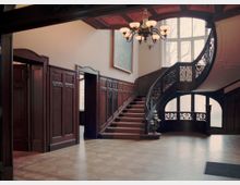 Historischer Innenraum mit Holzvertäfelung, einem geschwungenen Treppenaufgang mit verziertem Geländer und einem großen, bogenförmigen Fenster, das Tageslicht hereinlässt. Eine hängende Deckenlampe mit mehreren Leuchten und ein Bild an der Wand ergänzen die elegante Gestaltung.