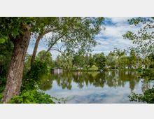 Seeufer mit Blick auf einen ruhigen See, umgeben von Bäumen und kleinen Häusern im Hintergrund, sowie einem Bootshaus am Ufer. Der Himmel ist leicht bewölkt, und das Wasser reflektiert die umliegende Vegetation.