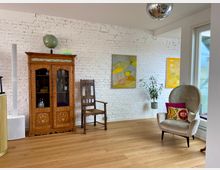 Ein helles Wohnzimmer mit weiß gestrichener Ziegelwand, einem Holzboden und moderner Möblierung. Zu sehen sind ein verziertes Holzvitrine, ein Polsterstuhl mit bunten Kissen, eine Zimmerpflanze, zwei Gemälde an der Wand und eine Discokugel an der Decke.