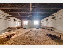 Ein leerer, verlassener Raum mit weiß verputzten, rissigen Wänden, zwei großen Fenstern mit Gitterstäben und einer Dachkonstruktion aus Metallträgern. Der Boden ist mit Sand bedeckt, während an den Seiten schmale Holzbänke und niedrige Holztische sichtbar sind.