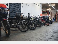 Reihe von Motorrädern verschiedener Marken und Modelle, die in einer Werkstatt mit grauem Boden und weißen Wänden abgestellt sind; im Hintergrund sind Regale, Leitern und Werkstattmaterialien sichtbar.
