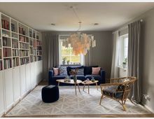 Wohnzimmer mit einem hellen, skandinavischen Design: Links eine weiße Bücherwand mit Regalen, in der Mitte ein blaues Sofa mit bunten Kissen vor einem großen Fenster mit grauen Vorhängen, daneben ein runder Holztisch auf einem weißen Teppich. Rechts steht ein Stuhl aus Rattan, und über dem Sofa hängt eine moderne Lampe mit Papieranhängern.