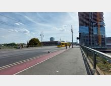 Straße mit Straßenbahn, Fahrradweg und Fußgängerweg auf einer Brücke. Im Hintergrund ein großes modernes Gebäude mit Glasfassade im Bau, eine Baumreihe und ein Kirchturm.