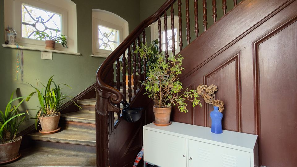 Treppenhaus mit geschwungener Holztreppe und dunklem Geländer, flankiert von zwei kleinen Fenstern mit dekorativem Gitter. Im Eingangsbereich stehen mehrere Topfpflanzen und ein weißes Sideboard vor einer holzvertäfelten Wand.