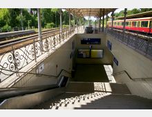 Bahnhof mit Treppenabgang zu einem unterirdischen Bereich, flankiert von Geländern mit filigranen Mustern. Im Hintergrund sind Gleise, ein rot-gelber Zug und grüne Vegetation zu sehen, während Beschilderungen und Infotafeln den Zugang zur S-Bahn kennzeichnen.