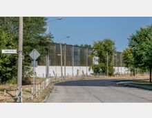 Straßenszene mit einer leichten Kurve, umgeben von Bäumen und Laternenmasten. Im Hintergrund steht eine große Industriehalle mit metallischen und teils durchscheinenden Fassadenelementen, daneben ein Straßenschild mit der Aufschrift „Schubertstraße“.