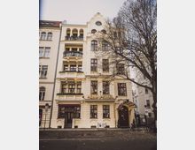 Fassade eines historischen Gebäudes mit dem Schriftzug "Henri Hotel" auf cremefarbener Fassade. Das Gebäude verfügt über mehrere Stockwerke, Balkone mit schmiedeeisernen Geländern und eine dekorative Giebelspitze; vor der Eingangstür ist ein Vordach mit geschwungenen Lampen angebracht, daneben stehen Fahrräder und ein Straßenbaum mit kahlen Ästen.