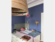Kinderzimmer mit blauer Wandfarbe, einem Bett mit grauem Kopfteil und eine Kommode mit Tischkicker-Spiel darauf. An der Wand hängen zwei dekorative Elefantenköpfe aus Naturmaterialien, und eine große, hängende Lampe ist im Vordergrund sichtbar. Die Decke hat Stuckverzierungen.
