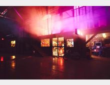 Industriehalle mit Treppe und großen Fenstern, erhellt durch rosa und orangefarbenes Licht. Im Erdgeschoss sind beleuchtete Werkstatträume mit sichtbaren Werkzeugen und Gegenständen, sowie ein geparktes Motorrad zu erkennen.