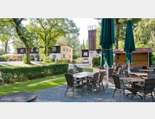 Außenbereich eines Cafés oder Restaurants mit Tischen und Stühlen auf einem Kiesboden, umgeben von grüner Vegetation und Bäumen, im Hintergrund ein mehrstöckiges Gebäude mit gelber und brauner Fassade sowie ein hoher Turm.